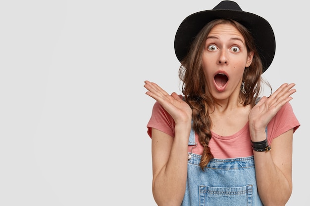 Mulher caucasiana surpresa e atônita vestida com macacão jeans, chapéu preto, mãos perto do rosto, encostada na parede branca com espaço livre para seu conteúdo promocional.