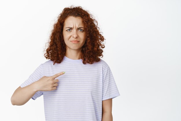 Mulher cética confusa apontando o dedo para si mesma, fazendo careta duvidosa, tem descrença em si mesma, fica contra o fundo branco.