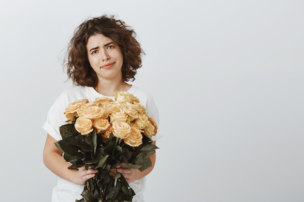 Mulher cética e sorridente segurando um buquê de rosas sem expressão impressionada