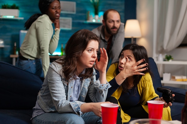 Mulher chateada depois de perder em videogames online enquanto se socializava com amigos multiétnicos na sala de casa tarde da noite, bebendo cerveja.