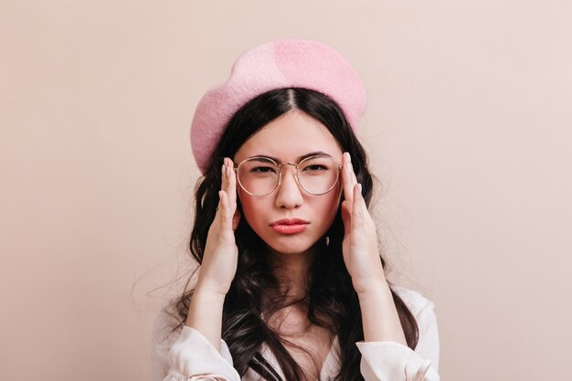 Mulher chinesa pensativa de óculos, olhando para a câmera. Modelo asiático engraçado na boina posando em fundo bege.