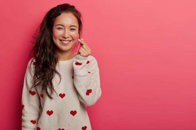 Mulher chinesa romântica mostra o signo do coração coreano com dois dedos cruzados, sorri com alegria e confessa que está apaixonada, expressa afeto, usa um suéter com estampa de coração, isolado na parede rosa do estúdio