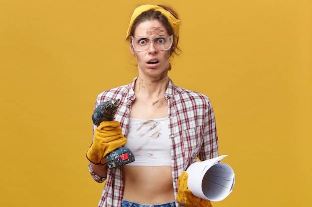 Foto grátis mulher chocada com luvas de proteção, camisa xadrez e tiara amarela segurando a broca e papel enrolado com o rosto sujo fazendo reparos dentro de casa ficando surpresa ao ver o quanto ela deve fazer