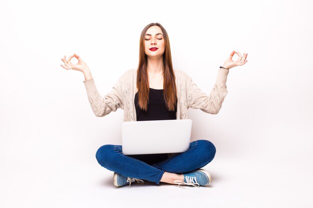 Mulher chocada e confusa em uma camiseta sentada no chão com um laptop, segurando óculos e olhando para a câmera sobre o cinza