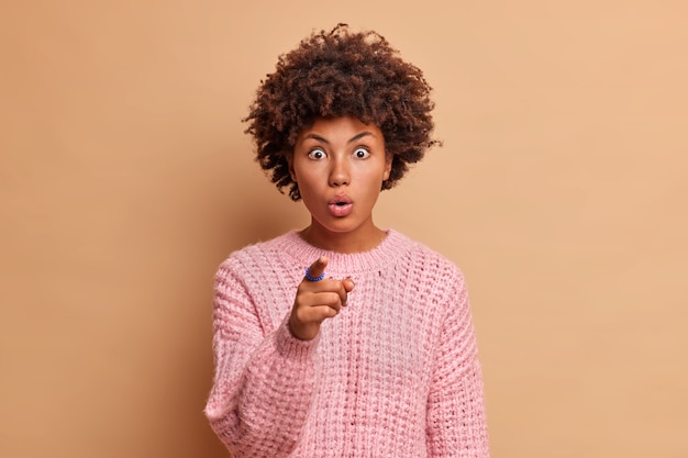 Mulher chocada e impressionada com cabelo encaracolado parece com grandes pontos de admiração diretamente na frente vê algo inacreditável poses sem palavras em ambientes internos vestida com malhas isoladas sobre parede bege