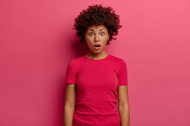 Mulher chocada e muda engasga de medo, percebe que esqueceu o passaporte enquanto chega ao aeroporto, fica atordoada e impressionada, usa uma camiseta casual, posa sobre a parede rosa, olha fixamente