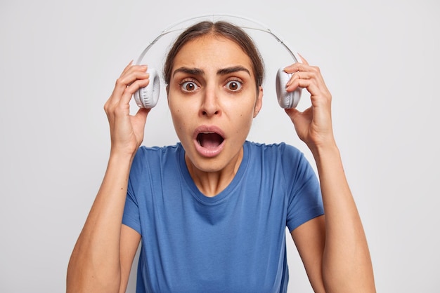 Mulher chocada tira os fones de ouvido ouviu algo com som muito alto hods respira mantém a boca aberta descobre algo incrível usa camiseta azul casual isolada sobre parede branca