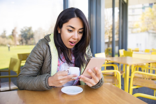 Mulher chocada usando smartphone e tomando café no café