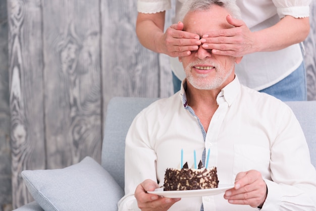 Mulher, cobertura, dela, marido's, olhos, segurando, bolo aniversário, em, mão