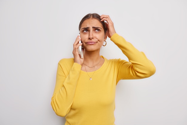 mulher coça a cabeça considera que algo se sente infeliz faz uma ligação mantém o celular perto da orelha vestida com um macacão amarelo casual sobre branco