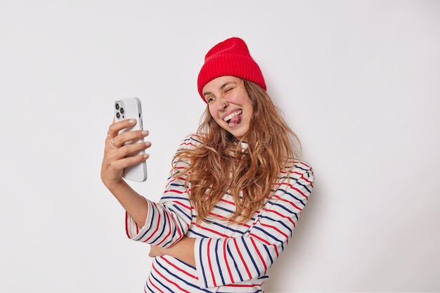 Mulher coloca a língua para fora faz uma careta engraçada enquanto toma selfie em smartphone vestido com roupa casual, isolado no branco. adolescente faz videochamada.