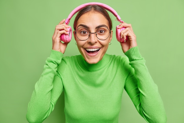 Foto grátis mulher coloca fones de ouvido, sorri alegremente ouve faixa de áudio está alegre, usa óculos redondos e gola olímpica no verde