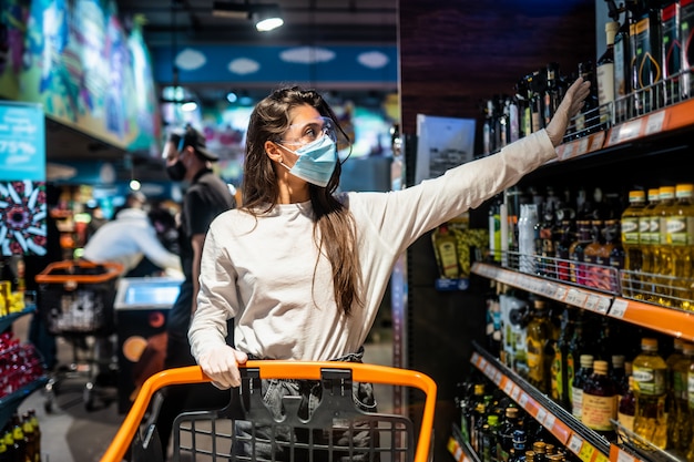 Mulher com a máscara cirúrgica e as luvas está fazendo compras no supermercado após a pandemia do coronavírus. A menina com máscara cirúrgica vai comprar a comida.