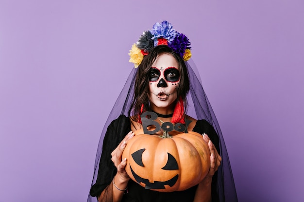 Mulher com arte facial em estilo mexicano está tentando assustar. Morena com abóbora e véu de casamento preto posando na parede lilás.