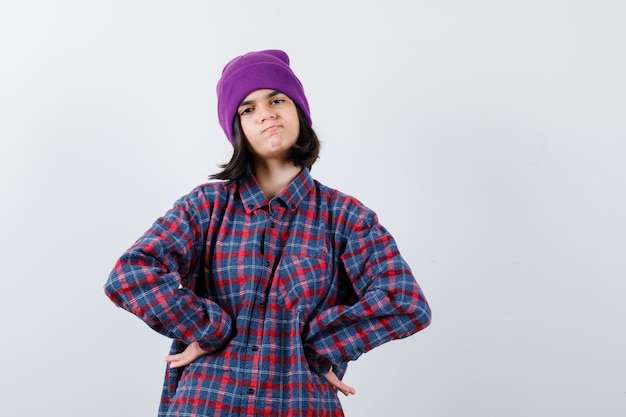 Mulher com as mãos no quadril com uma camisa quadriculada e um gorro parecendo melancólica