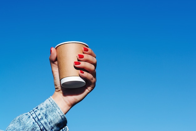 Mulher com as mãos segurando uma xícara de café sobre o fundo do céu azul. menina hippie segurando o copo de papel com café para levar. xícara de café de papel nas mãos da mulher com manicure perfeita.