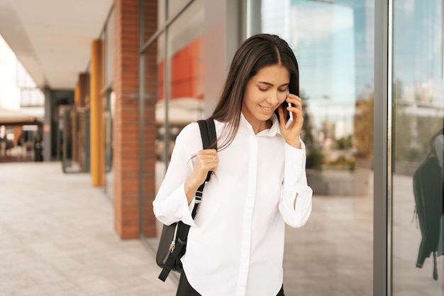 Mulher com backpak de negócios caminhando enquanto fala ao telefone perto de um prédio comercial