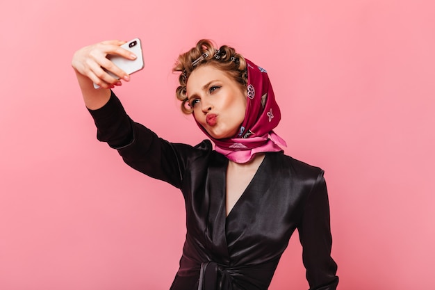 mulher com blusa de seda e lenço mandando beijo e fazendo selfie na parede rosa