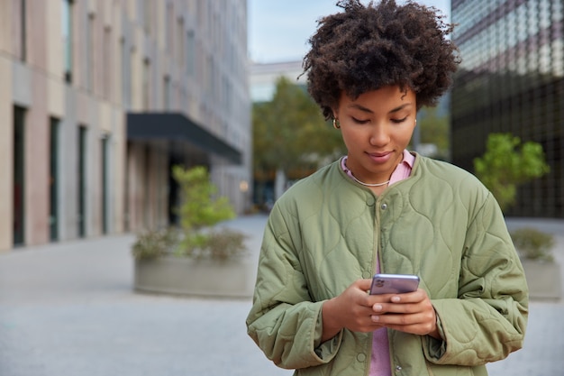 Mulher com cabelo afro segura um gadget de smartphone moderno para networking vestido de anoraque casual navega na internet lê boas notícias edita arquivos de mídia faz banco on-line