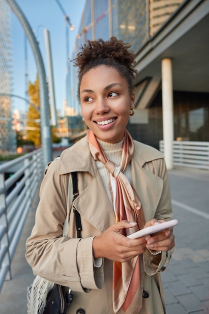 mulher com cabelo afro sorriso amplamente vestida com casaco bege passeia pela cidade durante o dia ensolarado de outono usa smartphone com prazer e se diverte enviando mensagens de texto no celular navega em páginas da web