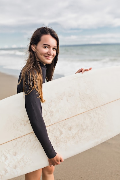 Mulher com cabelo comprido molhado posando para a câmera com uma prancha de surf perto do mar sob a luz do sol