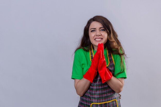 mulher com cabelo comprido ondulado usando avental e luvas de borracha implorando e rezando com as mãos juntas com expressão de esperança no rosto em pé