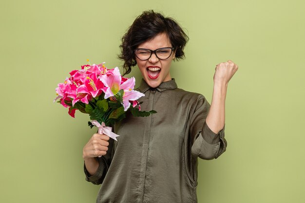 Mulher com cabelo curto segurando um buquê de flores olhando para a câmera feliz e animada com o punho cerrado celebrando o dia internacional da mulher, 8 de março, em pé sobre um fundo verde