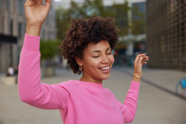 mulher com cabelo encaracolado dançando despreocupada, mantendo os braços levantados, vestida com um poloneck rosa casual fecha os olhos em poses de satisfação do lado de fora