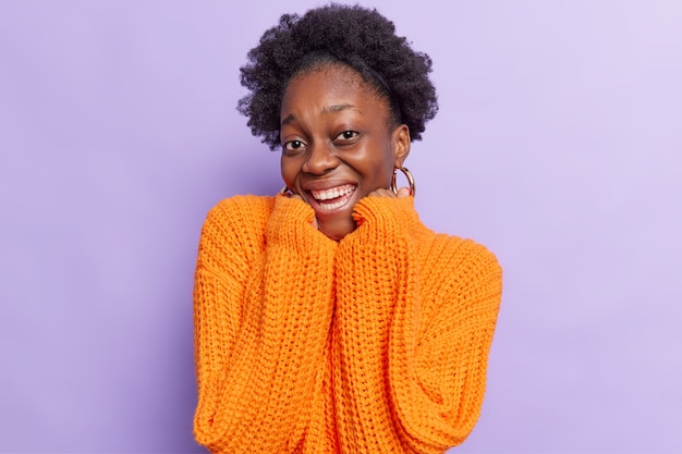 Foto grátis mulher com cabelo encaracolado mantém as mãos embaixo do queixo sorri positivamente mostra dentes brancos e de bom humor usa suéter laranja de malha tamanho isolado no roxo