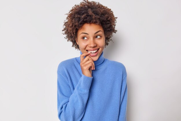 mulher com cabelo encaracolado mantém o dedo perto da boca sorri com os dentes olhando com admiração ao lado se sente tímida na presença de um estranho usa poses casuais de macacão azul em branco