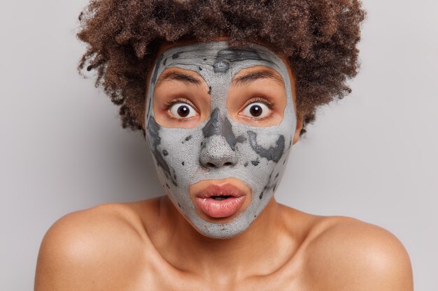 mulher com cabelo encaracolado olha impressionada aplica máscara facial de argila cuida de arfadas de pele seca de maravilha passa por tratamentos de beleza isolado no branco