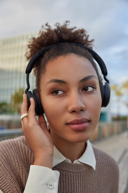 Mulher com cabelo encaracolado ouve música ou transmissão de rádio por meio de fones de ouvido sem fio aprecia a nova lista de reprodução usa equipamento estéreo moderno focado em algum lugar ao ar livre