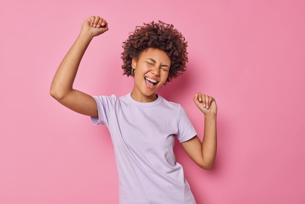 mulher com cabelo encaracolado se diverte e dança despreocupada vestida casualmente isolada na rosa gosta de festa e música favorita isolada na rosa. Pessoas e emoções
