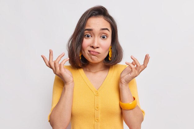 mulher com cabelo escuro expressão sem noção levanta as mãos encolhe os ombros com perplexidade Não sabe como agir usa um macacão amarelo casual isolado no branco