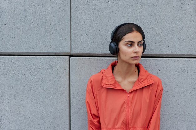 mulher com cabelo escuro vestida com um anoraque casual desvia o olhar e escuta música com fones de ouvido sem fio poses contra uma parede cinza espaço em branco ao ar livre para sua informação