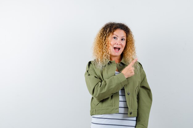 Mulher com cabelo loiro encaracolado, apontando para o canto superior direito com jaqueta verde e olhando feliz, vista frontal.