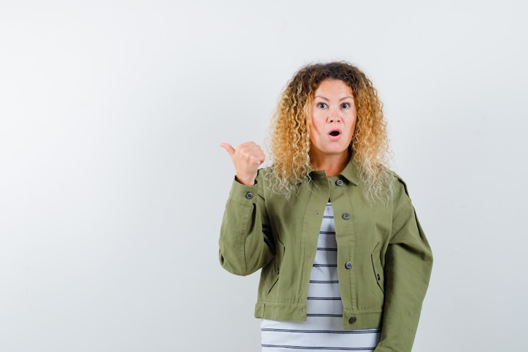 Foto grátis mulher com cabelo loiro encaracolado numa jaqueta verde apontando para o lado com o polegar e olhando maravilhada, vista frontal.
