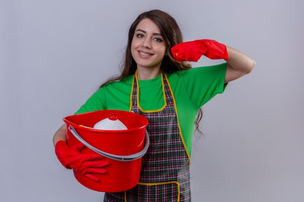 mulher com cabelo longo ondulado no avental e luvas segurando um balde com ferramentas de limpeza levantando o punho como uma vencedora sorrindo alegre em pé