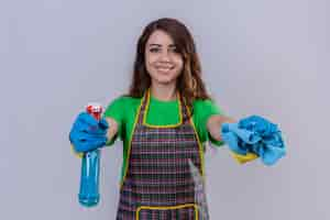 Foto grátis mulher com cabelo longo ondulado usando avental e luvas de borracha segurando um tapete e spray de limpeza, parecendo confiante e sorridente pronta para limpar em pé