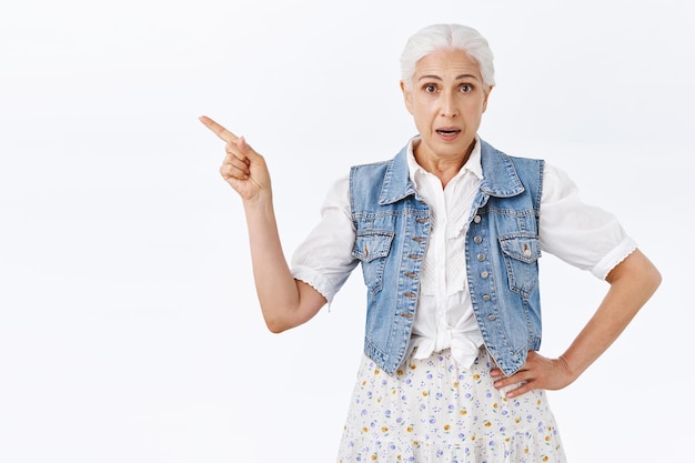 Mulher com cabelos grisalhos penteados, usar colete e vestido, apontar o dedo para a esquerda, fazer perguntas, consultar o balconista, comprar algo na loja