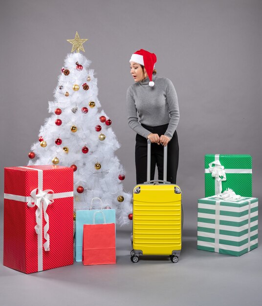 Mulher com chapéu de Papai Noel com bagagem ao lado da árvore de Natal