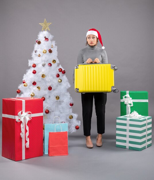 Mulher com chapéu de Papai Noel com bagagem ao lado da árvore de Natal