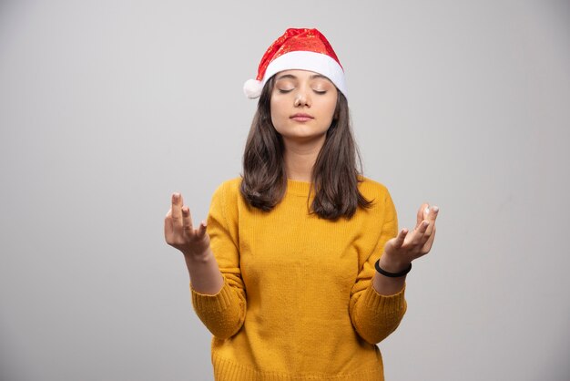 Mulher com chapéu de Papai Noel, fazendo meditação na parede cinza.