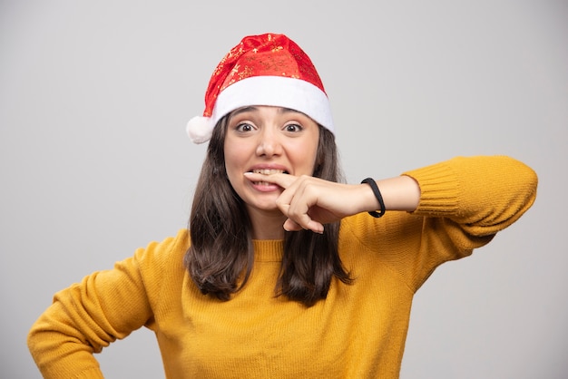 Mulher com chapéu de Papai Noel, mordendo o dedo na parede cinza.