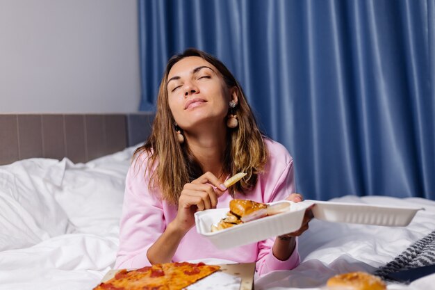 Mulher com comida rápida do leste desde a entrega na cama no quarto em casa Mulher desfrutando de pizza e hambúrgueres com comida gorda Com fome de carboidratos