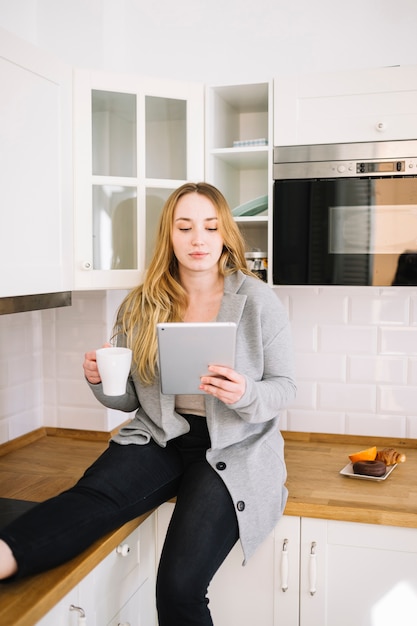 Mulher com copo usando tablet na cozinha