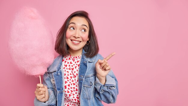 mulher com expressão alegre sorrindo agradavelmente indica distância no espaço da cópia mostra direção segura delicioso algodão doce usa jaqueta jeans