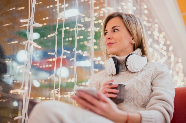 Mulher com fones de ouvido no pescoço e copo perto de luzes de Natal