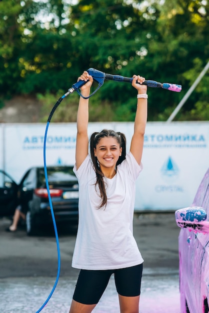 Foto grátis mulher com mangueira de alta pressão fica ao lado de um carro coberto de espuma rosa