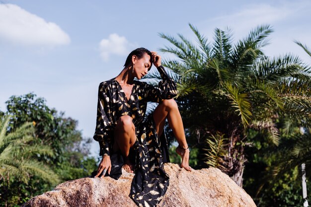 mulher com maquiagem de bronze em vestido preto dourado sentada na pedra